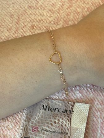 Minimalist delicate gold open-heart chain bracelet on a wrist atop a pink textured towel beside an aftercare card