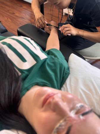 Nail salon scene — technician uses pink-handled cuticle nipper to groom a client's hand on a black armrest while the client relaxes in a chair.