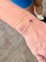 Close-up of a wrist wearing two dainty gold bracelets — a textured chain and a paperclip chain with a small crescent-moon charm — over a carpeted indoor floor.