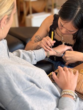 Close-up of a tattoo artist focused on inking a small wrist tattoo on a seated client in a cozy studio, pen-style tool and clamp visible with gold watch and bracelets on the wrist.