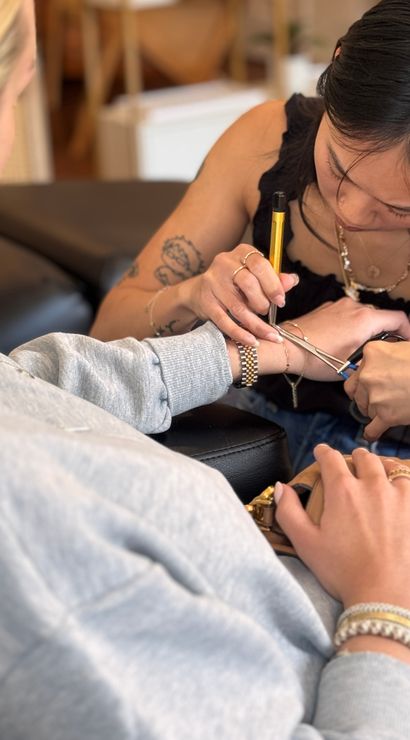 Close-up of a tattoo artist focused on inking a small wrist tattoo on a seated client in a cozy studio, pen-style tool and clamp visible with gold watch and bracelets on the wrist.