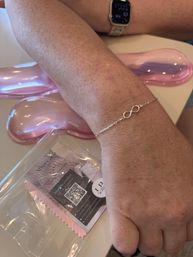 Close-up of a freckled forearm and hand wearing a delicate silver infinity bracelet and smartwatch, resting on a desk with pink gel wrist supports and a packaged jewelry cleaning cloth with a QR-code card.