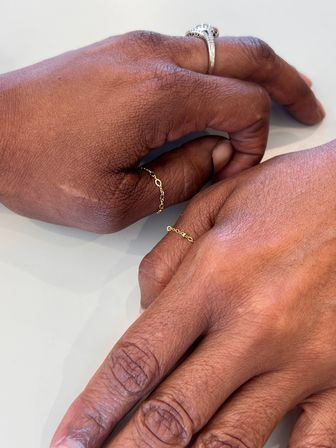Close-up of two brown hands on a light surface wearing delicate gold chain midi rings and a sparkling diamond engagement ring, minimalist jewelry with natural skin texture visible.