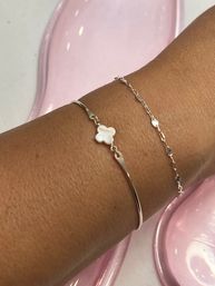 Close-up of a tanned wrist wearing two delicate silver-tone bracelets — a slim bangle with a white mother-of-pearl clover charm and a fine chain with tiny floral discs on a glossy pink background
