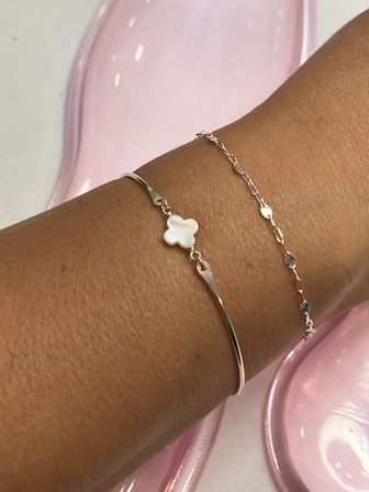 Close-up of a tanned wrist wearing two delicate silver-tone bracelets — a slim bangle with a white mother-of-pearl clover charm and a fine chain with tiny floral discs on a glossy pink background