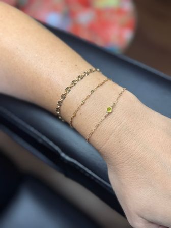 Close-up of a wrist wearing three delicate gold bracelets: a textured link bracelet, a thin chain with tiny floral discs, and a fine chain featuring a small yellow gemstone.