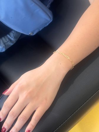 Close-up of a person's hand resting on black leather, showcasing a delicate gold chain-link bracelet, red painted nails and a blue purse in the background.