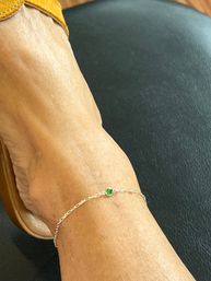 Close-up of a bare ankle wearing a delicate silver chain anklet with a small round green gemstone charm, tan sandal strap visible, foot resting on black leather seat — casual summer jewelry