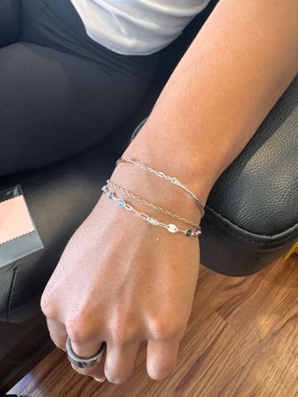Close-up of a wrist with stacked silver bracelets and a chunky silver ring, hand resting on a black leather chair over a wooden floor in an indoor setting.