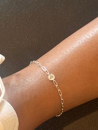 Delicate gold chain anklet with small round 'G' initial charm on a tanned ankle, close-up over a brown leather background.