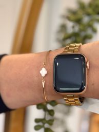 Close-up of a rose-gold square smartwatch with metal link band worn on a wrist next to a delicate gold bangle featuring a small white clover charm, stylish tech-meets-jewelry look