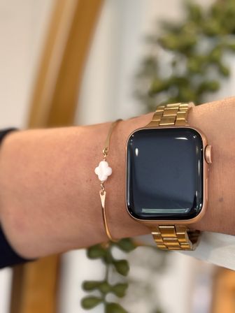 Close-up of a rose-gold square smartwatch with metal link band worn on a wrist next to a delicate gold bangle featuring a small white clover charm, stylish tech-meets-jewelry look
