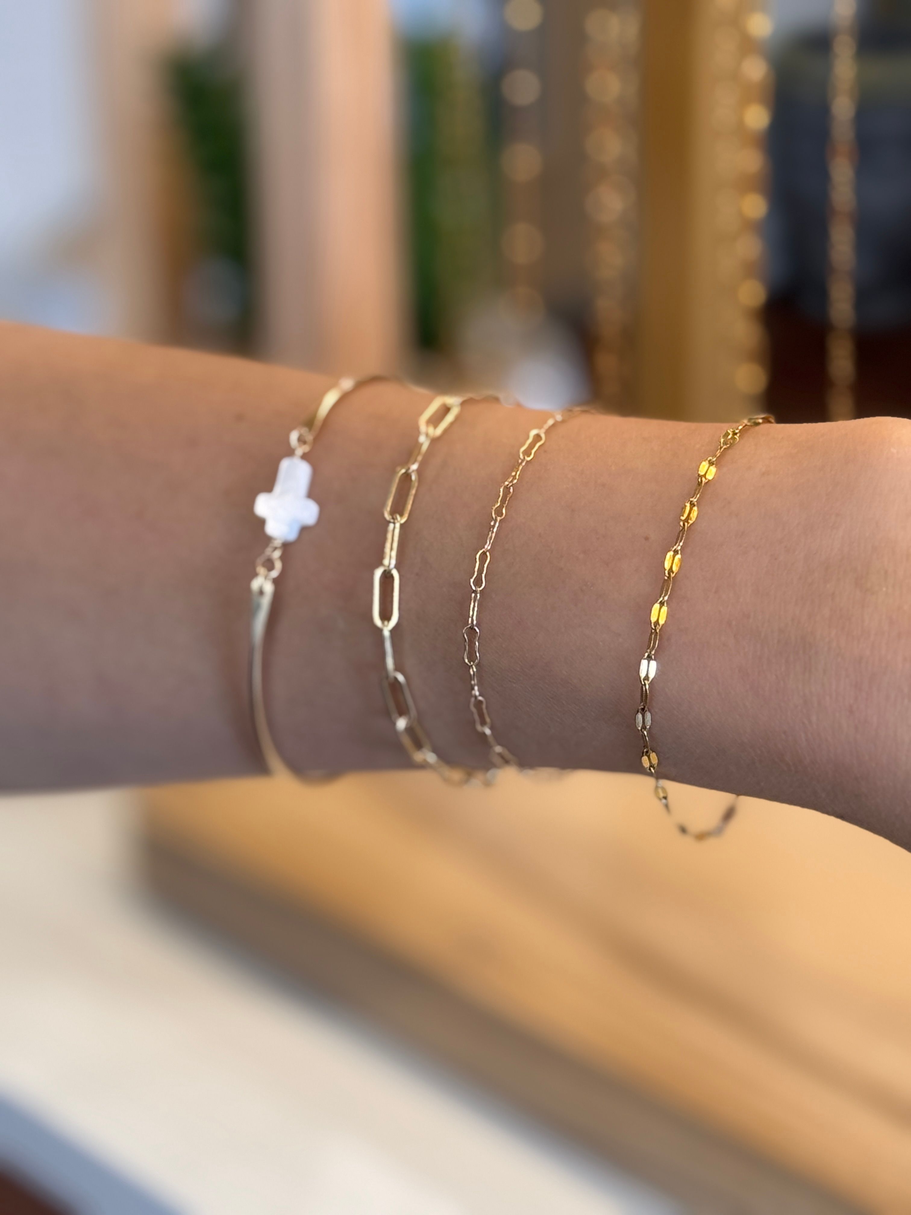 Stacked delicate gold bracelets on a wrist — thin bar, paperclip and fine chains, beaded link and a small white cross charm against a blurred boutique background