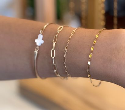 Stacked delicate gold bracelets on a wrist — thin bar, paperclip and fine chains, beaded link and a small white cross charm against a blurred boutique background