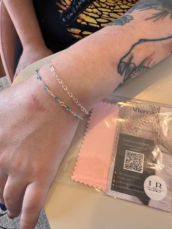 Close-up of a tattooed forearm wearing two delicate silver bracelets—one with small turquoise beads—resting beside a pink jewelry cleaning cloth and an aftercare card on a tabletop.