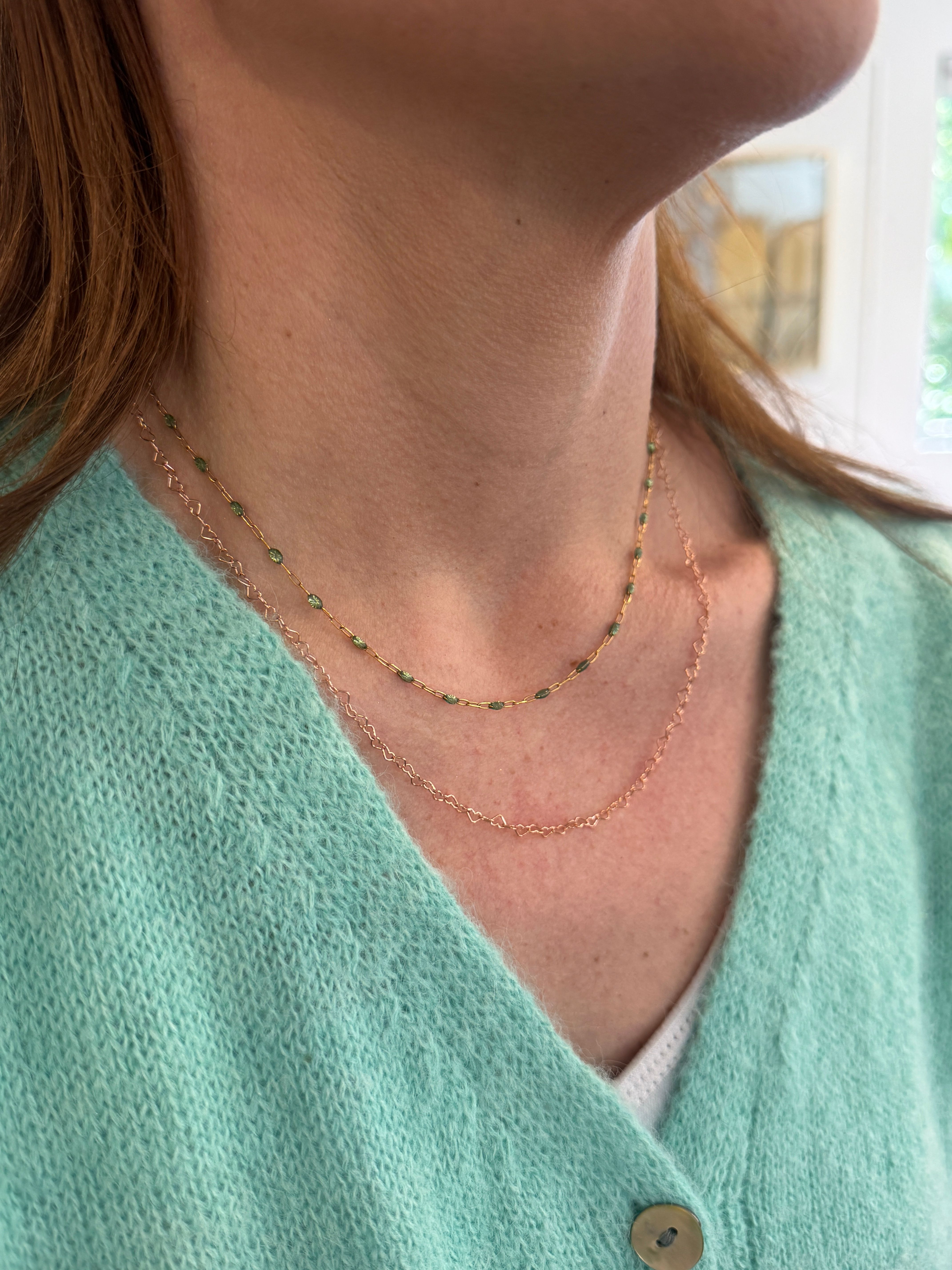 Close-up of a neckline wearing two delicate gold necklaces — one with tiny green beads and one fine twisted chain — paired with a mint-green knit sweater and visible button.