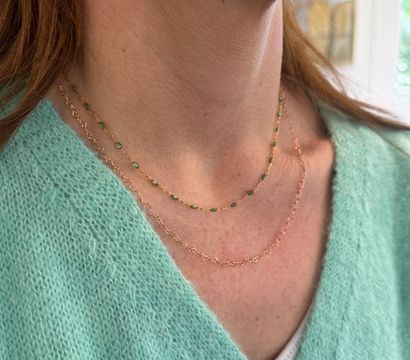 Close-up of a neckline wearing two delicate gold necklaces — one with tiny green beads and one fine twisted chain — paired with a mint-green knit sweater and visible button.