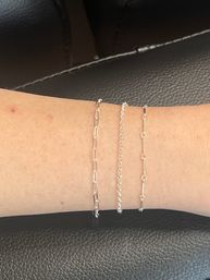 Close-up of three delicate stacked gold chain bracelets with different link styles on a forearm against black textured leather background — minimalist women's jewelry.