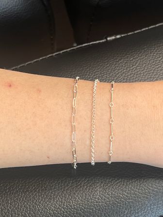 Close-up of three delicate stacked gold chain bracelets with different link styles on a forearm against black textured leather background — minimalist women's jewelry.