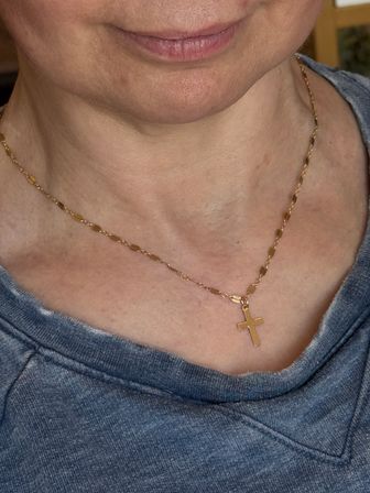 Close-up of lower face and neck wearing a shiny gold cross pendant on a delicate chain over a blue-gray sweatshirt.