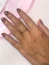 Close-up of hand with glossy taupe manicure wearing a delicate gold ring set with a small green gemstone, resting on a pink dish over a white surface.