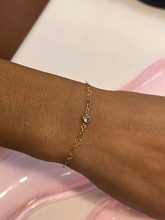 Close-up of a wrist wearing a dainty gold chain bracelet with a small bezel-set clear crystal and tiny star charm against a glossy pink background