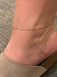 Close-up of a foot and ankle wearing a delicate gold chain anklet with tiny bead accents above a beige suede mule shoe on a gray carpeted floor.