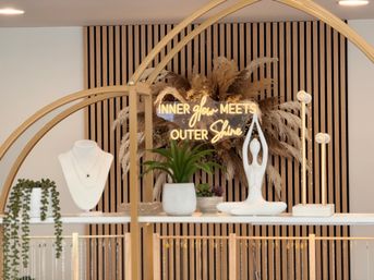 Modern retail display with neon sign reading 'INNER glow MEETS OUTER Shine', pampas grass and vertical wood slat backdrop, white jewelry bust, potted succulents and a white yoga sculpture on gold-framed shelving.