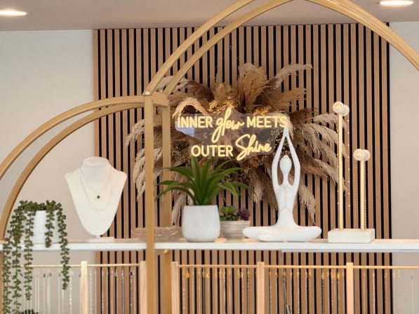 Modern retail display with neon sign reading 'INNER glow MEETS OUTER Shine', pampas grass and vertical wood slat backdrop, white jewelry bust, potted succulents and a white yoga sculpture on gold-framed shelving.