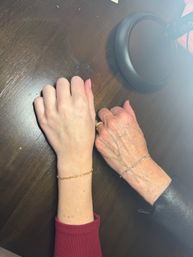 Close-up of two hands on a dark wood table — a younger hand with pink nails and a delicate gold chain bracelet holding an older hand wearing a silver bracelet and gold rings.