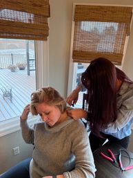 At-home haircut: seated woman in a gray sweater having her hair trimmed by another person beside a window with bamboo blinds, pink-handled hair tools on the table and a snowy backyard deck visible outside.