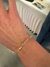 Close-up of a wrist wearing a delicate gold chain bracelet with curved script-like links, soft indoor background with a white tablecloth.