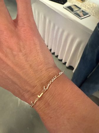 Close-up of a wrist wearing a delicate gold chain bracelet with curved script-like links, soft indoor background with a white tablecloth.