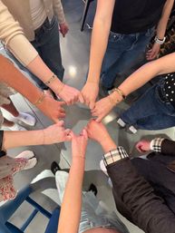 Overhead shot of friends standing in a circle linking pinkies, each wrist wearing delicate gold bracelets with casual jeans and socks on a polished gray floor — playful group jewelry moment.