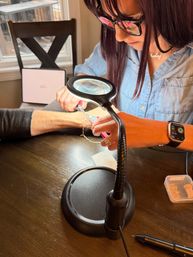 Close-up of jewelry repair: hands using pliers under a magnifying lamp to fix a silver bracelet on a wrist at a home workspace.