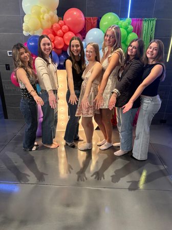 Seven friends in casual outfits smiling and extending their hands toward the camera in front of a colorful balloon arch and lighted marquee at an indoor party backdrop.
