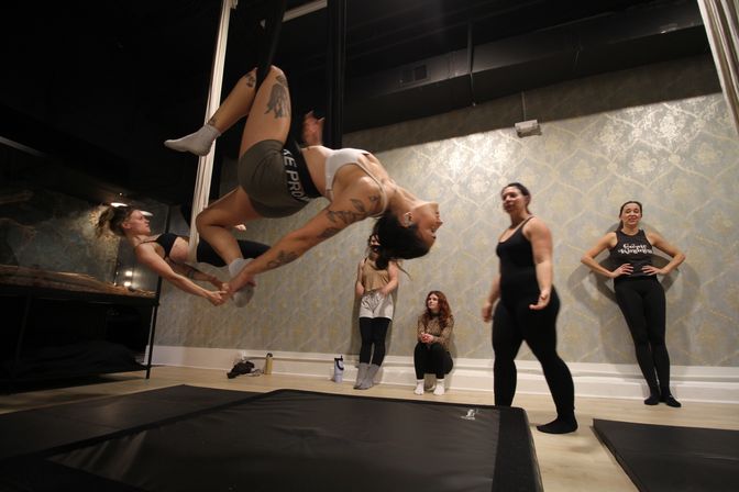 Aerial silks class in a fitness studio: two women suspended in an inverted partner pose while classmates and an instructor watch