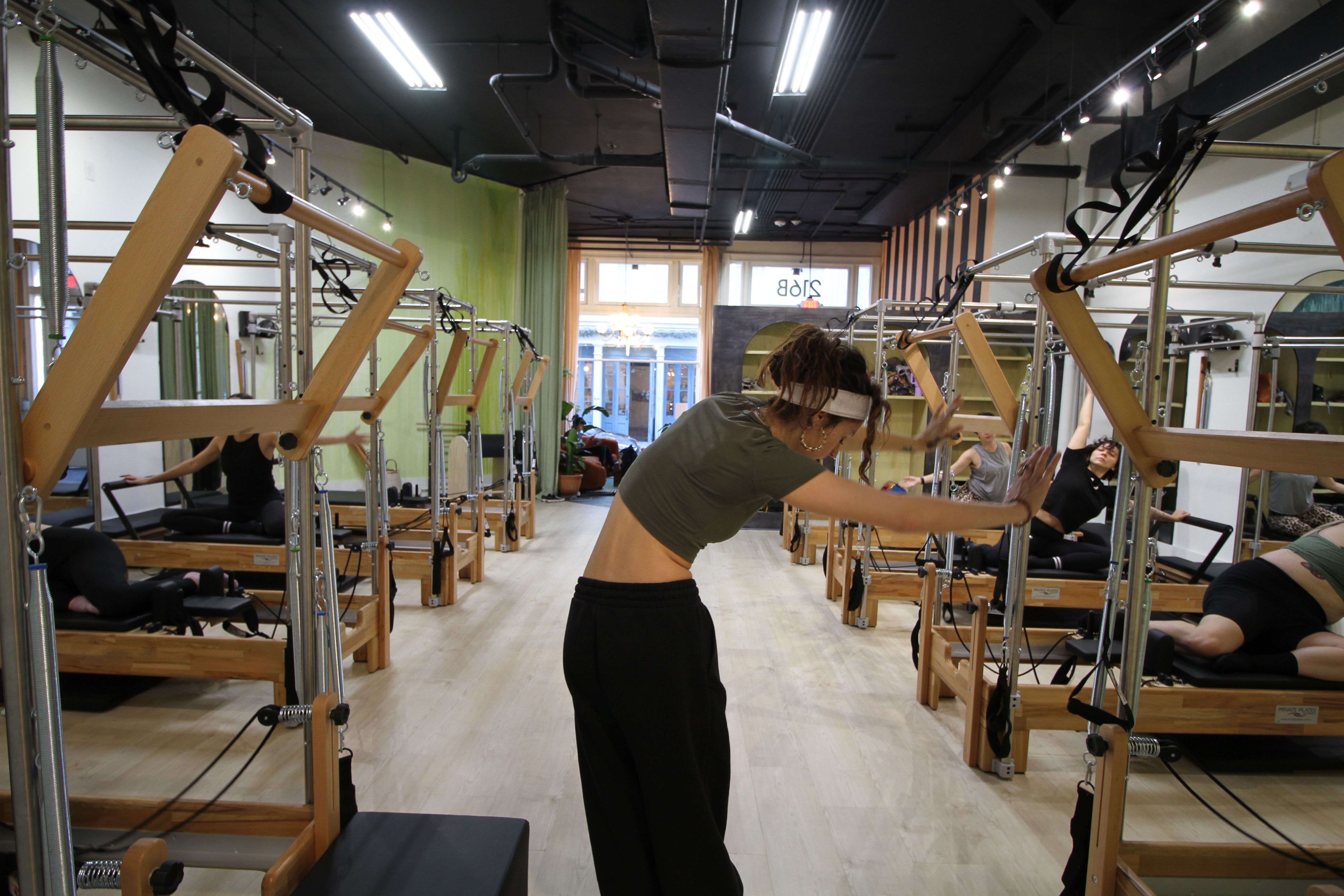 Bright urban pilates studio interior with rows of wooden reformer machines, light wood floors, and people stretching during a group class.