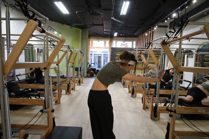 Bright urban pilates studio interior with rows of wooden reformer machines, light wood floors, and people stretching during a group class.