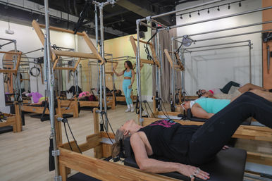 Pilates reformer class in a bright modern fitness studio — women doing core-strength exercises on wooden reformer machines while an instructor guides the session.