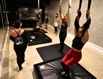 Guided group aerial yoga in an indoor fitness studio — women practicing on hanging silk hammocks over black padded mats on a light wood floor.