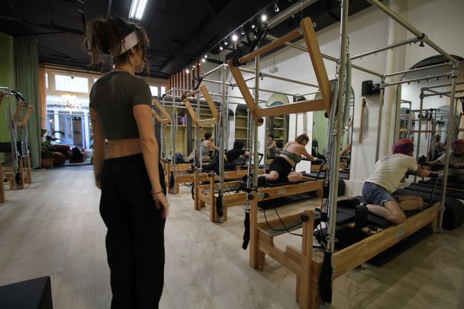 Group Pilates reformer class in a bright urban studio with wooden reformer machines and mirrors, instructor observing participants stretching.