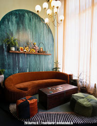 Cozy New Orleans French Quarter–style living room with a curved rust-orange velvet sofa, teal arched textured accent wall with shelf decor, vintage trunk coffee table, poufs, plants, and sheer floor-to-ceiling curtains.