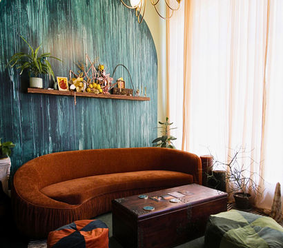 Cozy New Orleans French Quarter–style living room with a curved rust-orange velvet sofa, teal arched textured accent wall with shelf decor, vintage trunk coffee table, poufs, plants, and sheer floor-to-ceiling curtains.