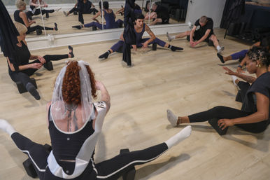 Group stretching class in a bright fitness studio — adults seated on yoga blocks doing wide-legged stretches around an aerial silk, mirrored wall reflecting the session.