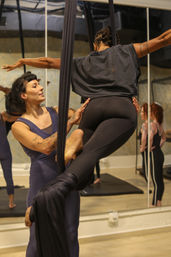 Aerial silks class in a mirrored fitness studio — instructor assists a student wrapped in black fabric as she balances in leggings and a t‑shirt, practicing aerial fitness.