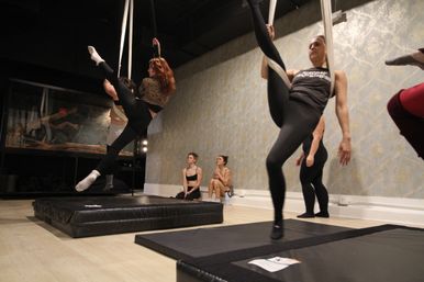 Aerial silks class in a dance studio — students practice high-leg holds and splits on white silks above padded crash mats