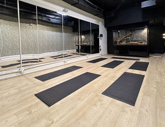 Minimal urban yoga studio interior with light wood flooring, mirrored wall and eight black yoga mats neatly spaced for class, dark back wall with a large terrarium display.