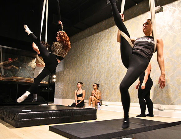 Group aerial silks class in an indoor fitness studio, women in leggings performing suspended leg extensions on white silks above padded mats