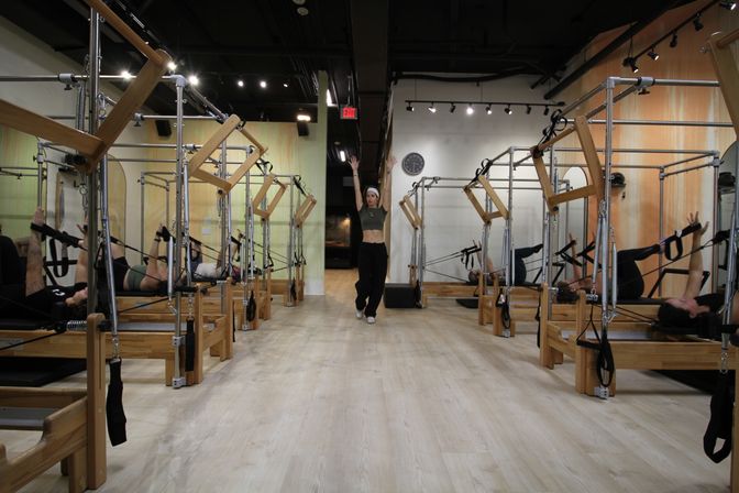 Modern Pilates studio with wooden reformer machines lining both sides, students using straps on the reformers, and an instructor standing in the center aisle with arms raised